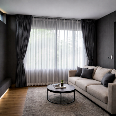 ห้องนั่งเล่นสไตล์มินิมอลที่มีโซฟาสีเบจ โต๊ะกาแฟสีดำทรงกลม ผ้าม่านจีบทึบสีเทาเข้มและผ้าโปร่งสีขาว และแสงธรรมชาติส่องผ่านหน้าต่างบานใหญ่