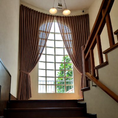 บันไดไม้ภายในบ้านที่มีหน้าต่างทรงโค้งติดผ้าม่านจีบสีน้ำตาลผูกเปิด มองเห็นวิวต้นไม้ด้านนอกและโคมไฟเพดานสามดวง