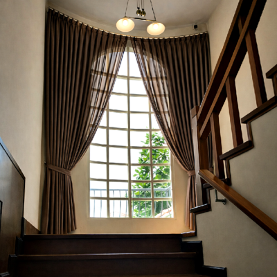 บันไดไม้ภายในบ้านที่มีหน้าต่างทรงโค้งติดผ้าม่านจีบผ้าโปร่งแสงสีน้ำตาลผูกเปิด มองเห็นต้นไม้ด้านนอกและโคมไฟเพดานสามดวง
